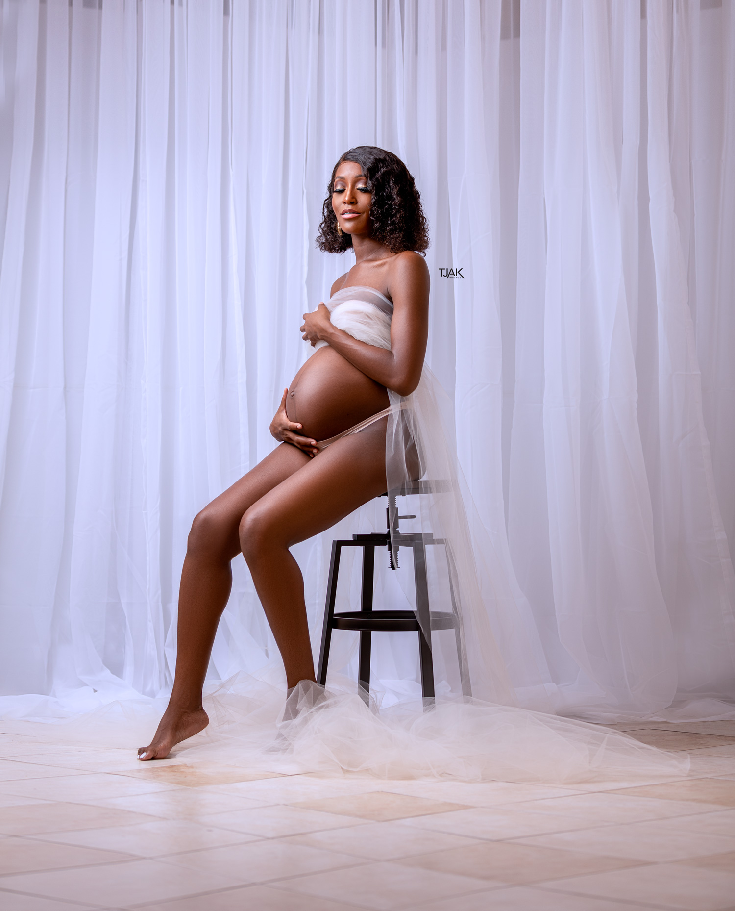 a maternity photograph of a pregnant woman sitting on a bar stool wrapped with a nude long tulle in a studio in Laurel MD near Baltimore Maryland in Howard County MD
